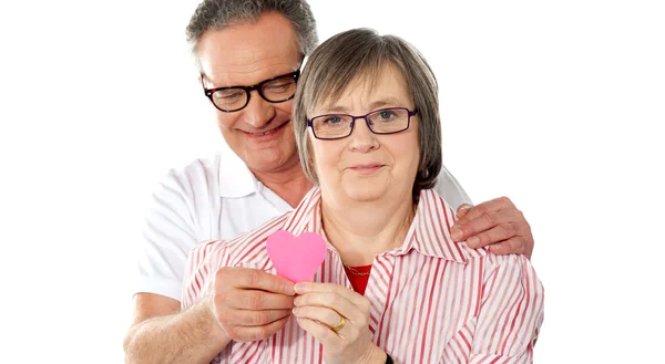 Old couple facing camera with focus on paper heart — Stock Photo, Image
