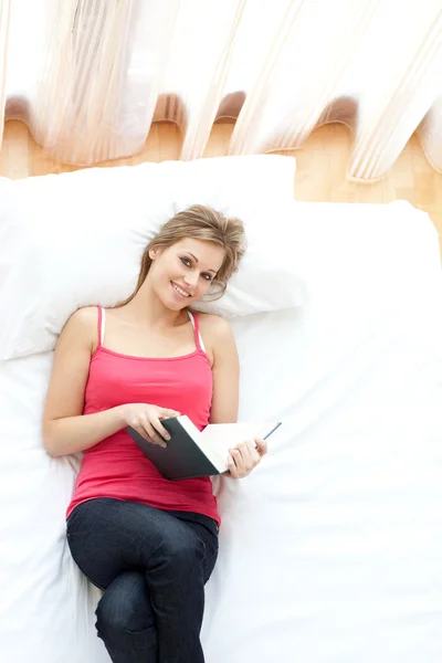 Leende kvinna läser en bok som ligger på sin säng — Stockfoto