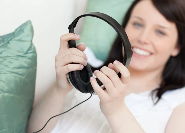 Χαμογελώντας νεαρή γυναίκα βάζει ακουστικά ξαπλωμένη σε ένα καναπέ — Φωτογραφία Αρχείου