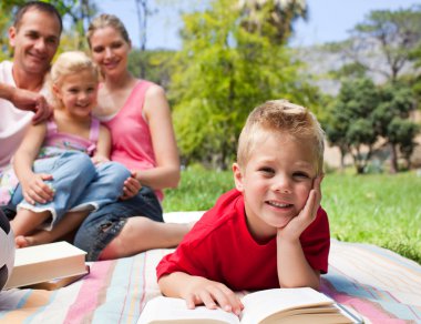 Smiling little boy reading lying on the grass while having a pic clipart