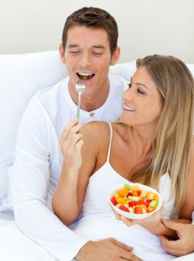 Jolly couple eating fruit lying on their bed clipart