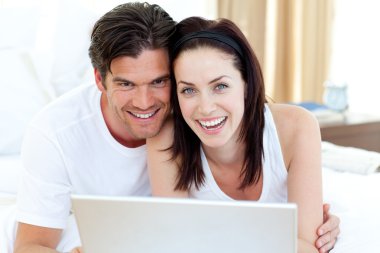 Smiling couple using a laptop lying on their bed clipart