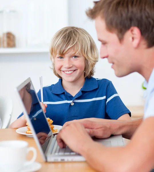 Schattige jongen ontbijten terwijl zijn vader met behulp van een laptop — Stockfoto