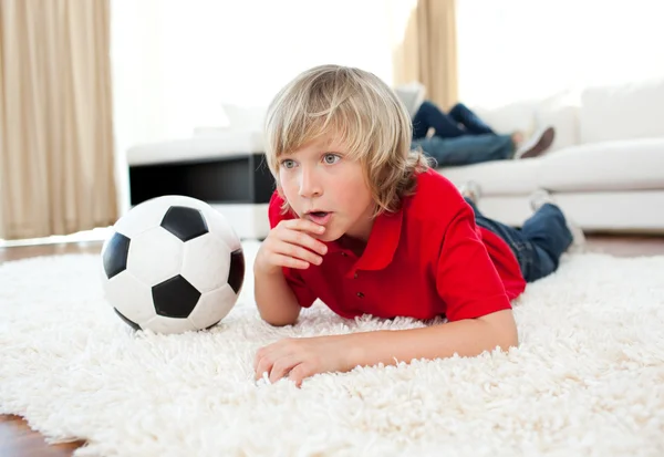 Menino animado assistindo jogo de futebol deitado no chão — Fotografia de Stock