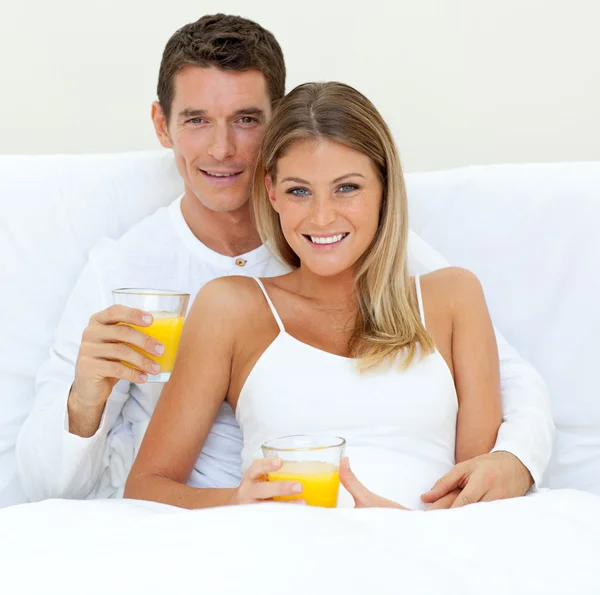 Ευτυχισμένο ζευγάρι πίνοντας χυμό πορτοκαλιού, ξαπλωμένος στο κρεβάτι τους — Φωτογραφία Αρχείου