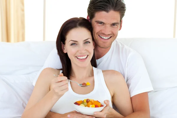 Happy couple eating fruits lying on their bed — Stock Photo, Image
