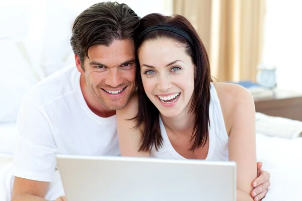 Smiling couple using a laptop lying on their bed — Stock Photo, Image