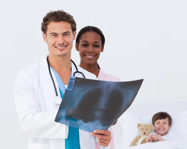 Улыбающийся доктор и медсестра осматривают рентген и ребенка в постели — стоковое фото