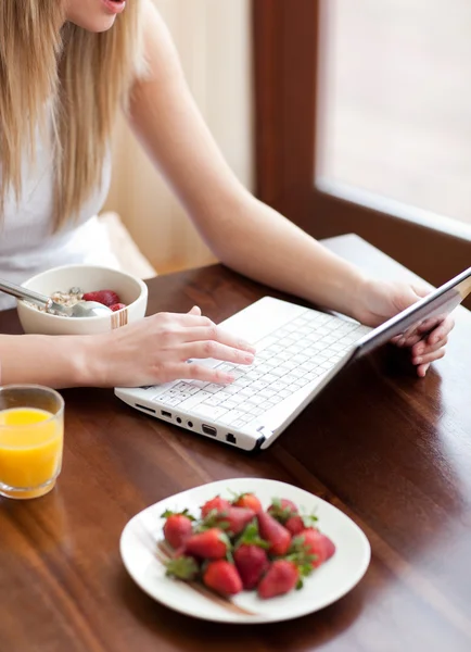 Sarışın bir kadın Kahvaltı yaparken bir dizüstü bilgisayar kullanarak — Stok fotoğraf