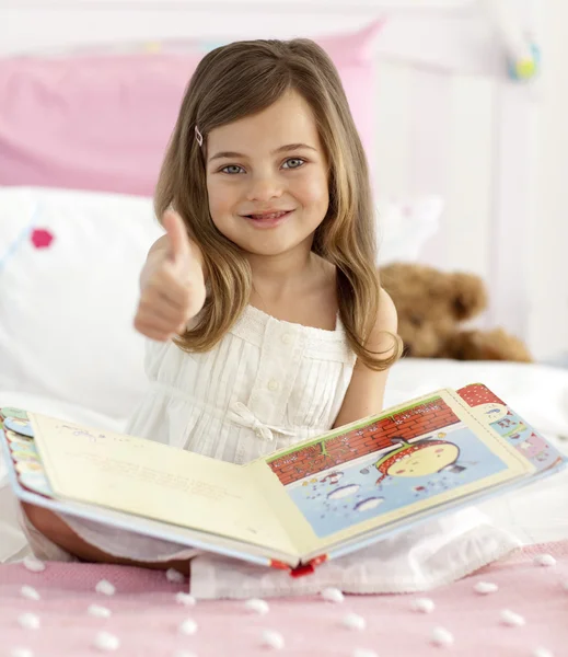 Gelukkig meisje lezen in bed met duim omhoog — Stockfoto
