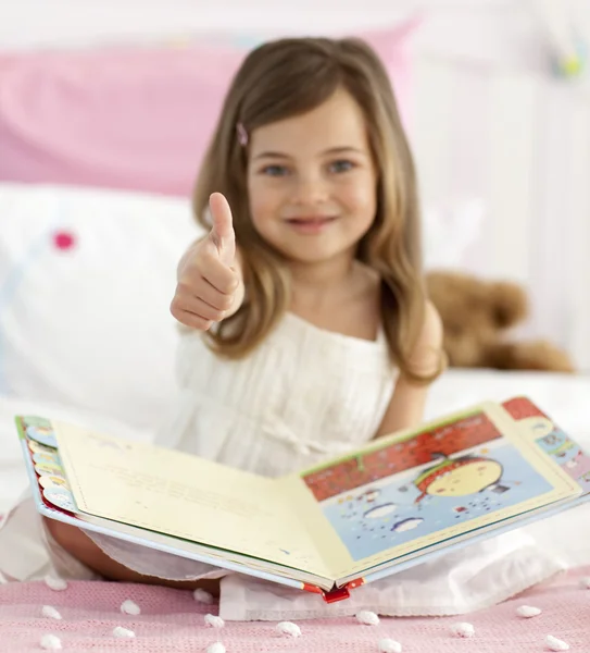 Маленькая девочка читает в постели с большим пальцем вверх — стоковое фото