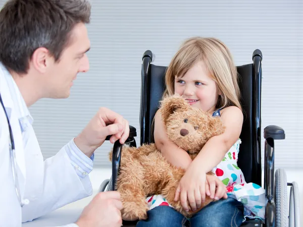 Улыбающаяся маленькая девочка, сидящая на инвалидном кресле и подглядывающая за доктором — стоковое фото