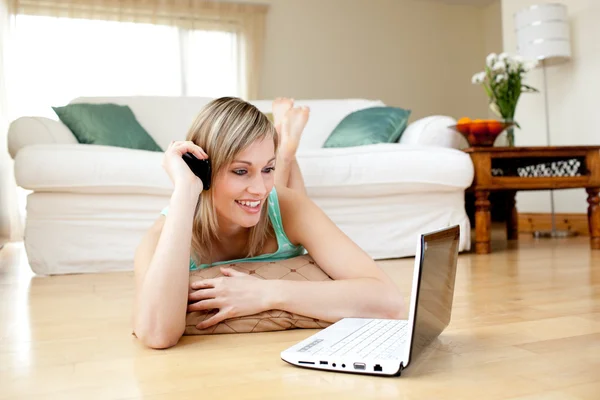 Mulher atraente no telefone ao usar um laptp deitado no chão — Fotografia de Stock