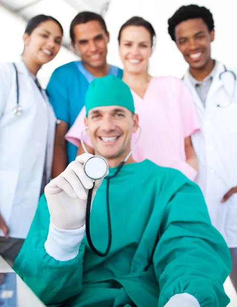 Cirujano sonriente sosteniendo un estetoscopio con su equipo en la espalda —  Fotos de Stock