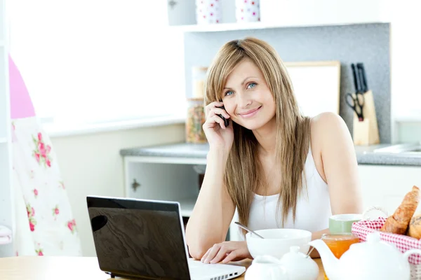 Mulher branca usando um laptop e um telefone na cozinha — Fotografia de Stock