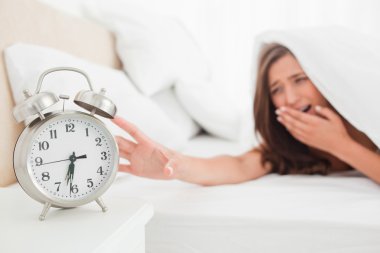 Woman yawning and reaching out for alarm clock from under her bl clipart