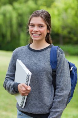 Smiling teenage girl holding a notebook while standing in a park clipart