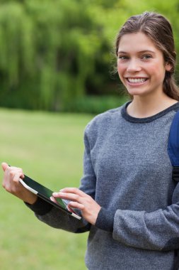 Smiling student using her tablet pc while standing in a park clipart