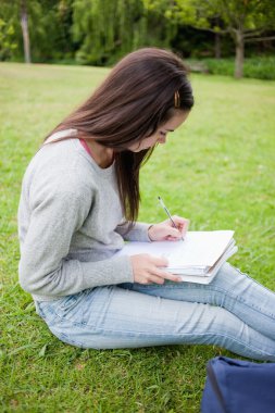 Young serious student sitting in a park while writing on a noteb clipart