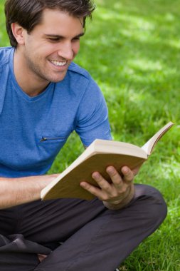 Young smiling man reading a book while siting on the grass clipart