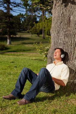 Man using headphones to sing along to music while resting a tree clipart