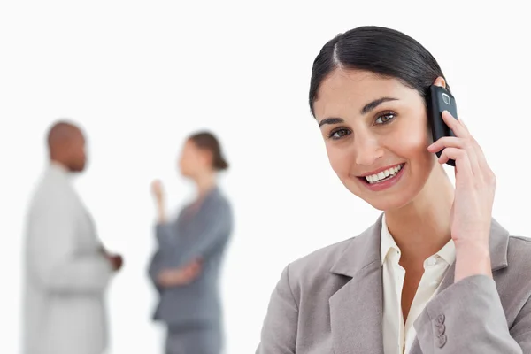 Smiling saleswoman with mobile phone and colleagues behind her — Stock Photo, Image