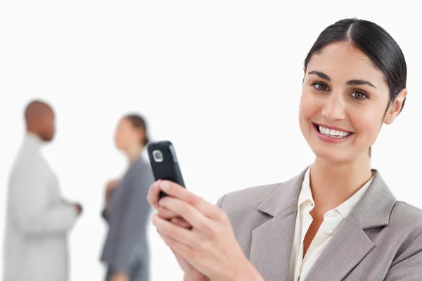 Χαμογελώντας πωλήτρια κρατώντας το κινητό τηλέφωνο με τους συναδέλφους πίσω της — Φωτογραφία Αρχείου