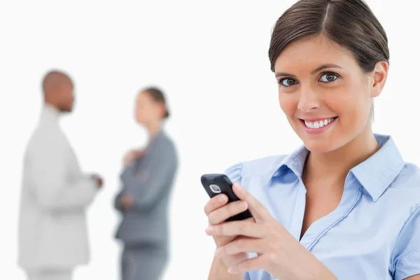 Vendedora sorrindo segurando telefone celular com colegas por trás h — Fotografia de Stock