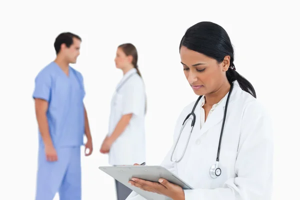 Doctor taking notes with talking staff members behind her — Stock Photo, Image