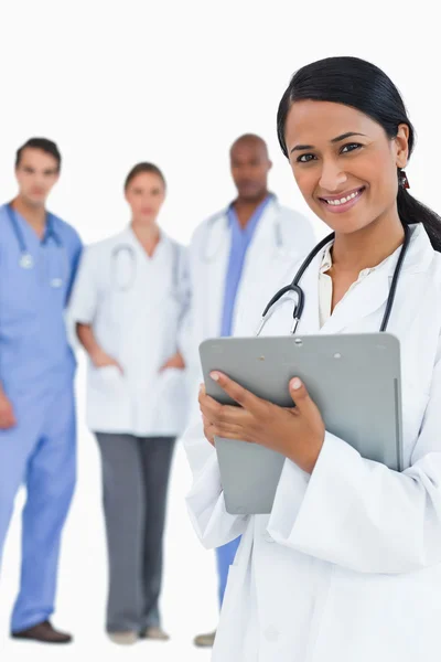 Smiling female doctor with clipboard and staff behind her — Stock Photo, Image