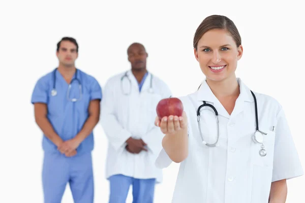 Médico oferecendo maçã com colegas por trás dela — Fotografia de Stock