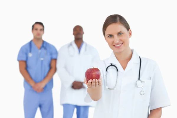 Dottore sorridente che offre mela con i colleghi dietro di lei — Foto Stock