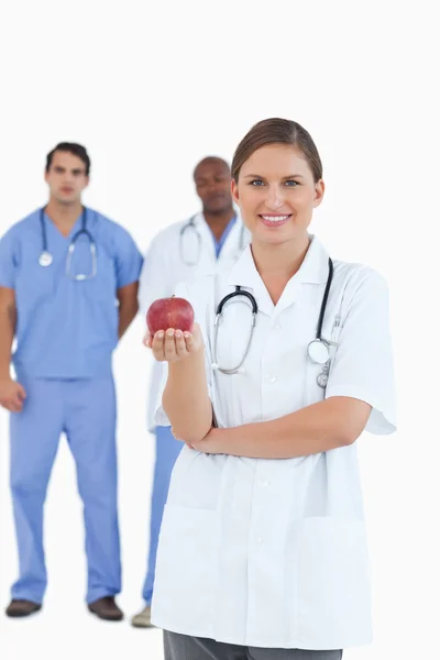 Médico sonriente con manzana y colegas detrás de ella — Foto de Stock