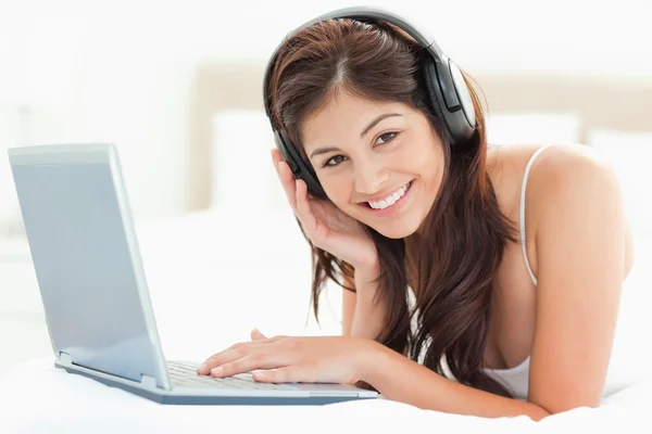 Woman lying on a bed using a laptop and listening to headphones — Stockfoto