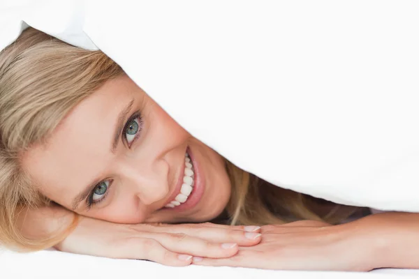 Женщина лежит под одеялом, прикрывая щеку, когда улыбается. — стоковое фото