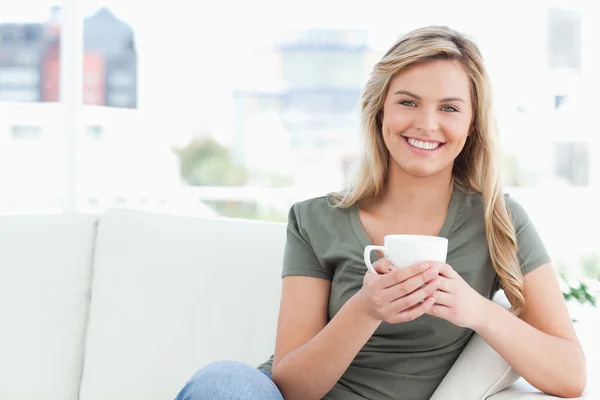 Mujer sonriendo, sosteniendo una taza en las manos y sentada en el sofá —  Fotos de Stock