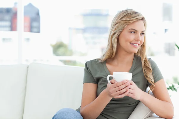 Woman holding a cup, sitting on the couch, looking sideways and — Stock Photo, Image