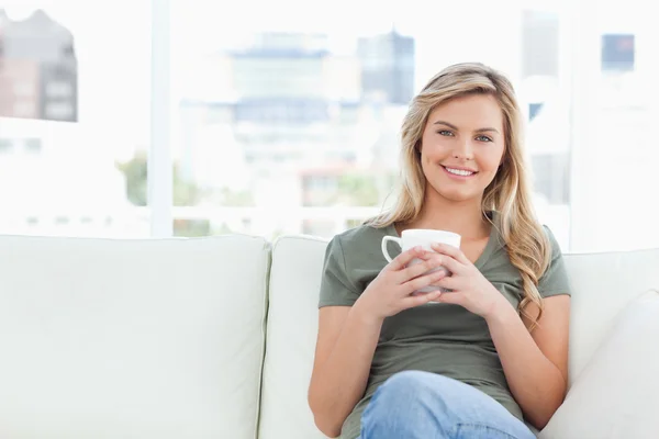 Woman sitting on couch, smiling and holding a cup in front of he — Stock Photo, Image