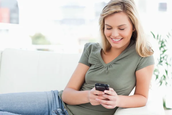 Frau lächelt, während sie ihr Handy benutzt und auf der Couch liegt — Stockfoto
