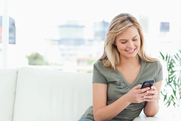 Žena sedí na gauči, pohledu a její telefon a s úsměvem — Stock fotografie