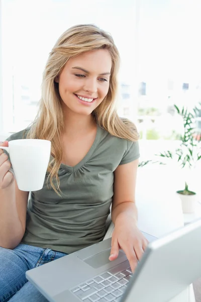 Donna sorridente come lei usa il suo computer portatile e tiene una tazza nel suo libero — Foto Stock
