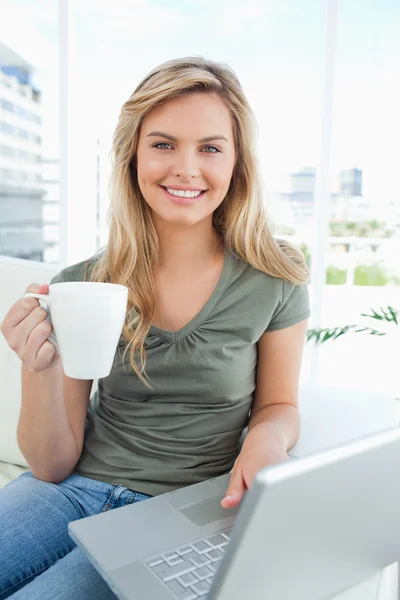 Woman looking forward with a cup in her hand, as she uses her la — Stock Photo, Image