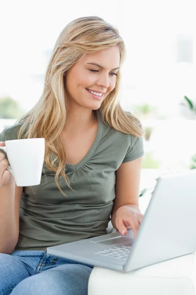 Woman smiling, as she uses her laptop and hold a cup in her othe — Stock Photo, Image