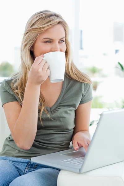 Woman taking a drink while using her laptop and looking to the s — Stock Photo, Image