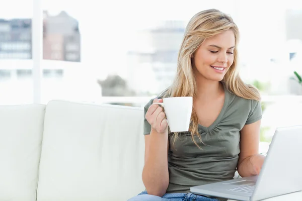Woman smiling as she holds her cup and uses her laptop — Stock Photo, Image