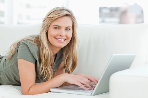 Frau liegt mit Laptop vor sich auf der Couch, lächelt — Stockfoto