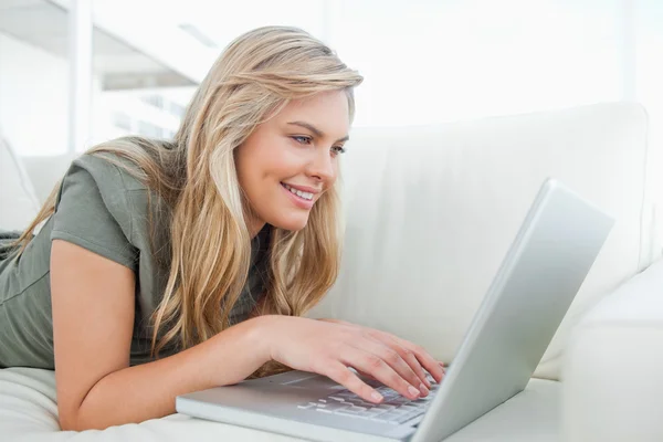 Mujer sonriendo mientras usa su portátil mientras está acostada en el sofá — Foto de Stock