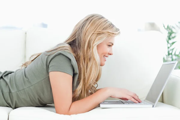 Mulher usando seu laptop e sorrindo enquanto ela se deita no sofá — Fotografia de Stock