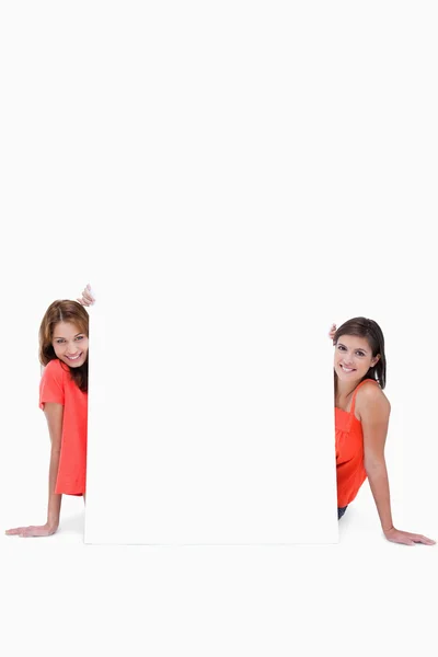 Two smiling teenage girls sitting behind a blank poster — Stock Photo, Image