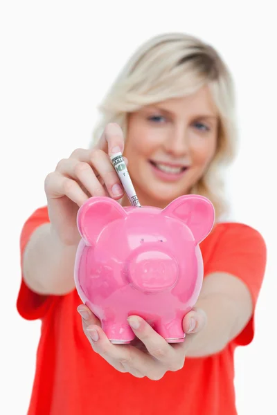 Jovem loira mulher colocando notas em um rosa porquinho banco — Fotografia de Stock